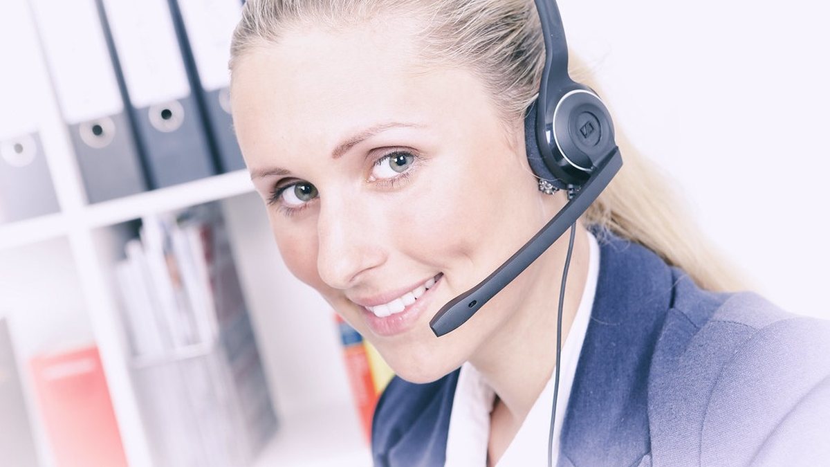 Nữ nhân viên đeo tai nghe headset mỉm cười, đang làm việc kiểu call center/tổng đài, phía sau là tủ hồ sơ văn phòng